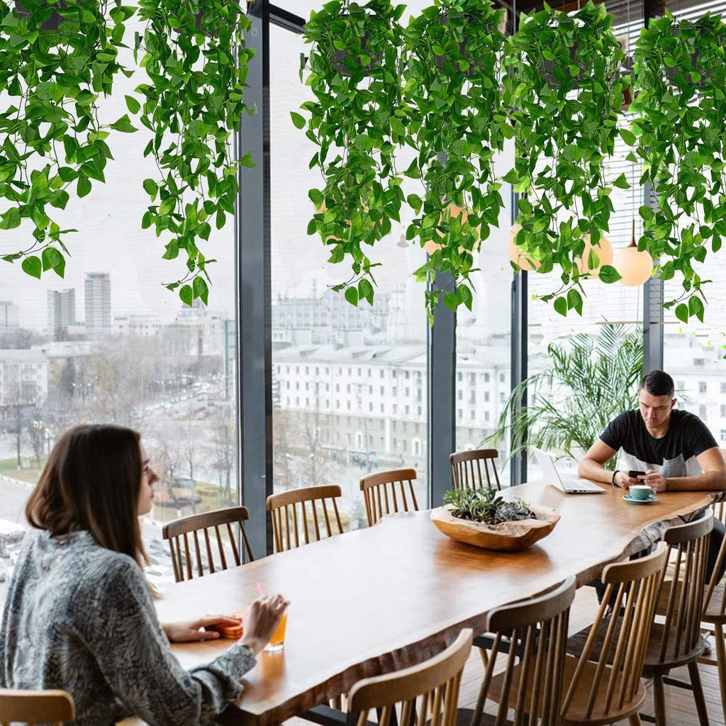 Two artificial hanging plants with green ivy vines draped over a large wooden table in a modern dining area with people seated.