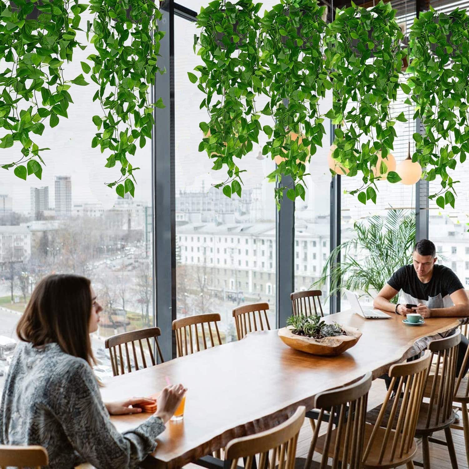 Two artificial hanging plants with green ivy vines draped over a large wooden table in a modern dining area with people seated.