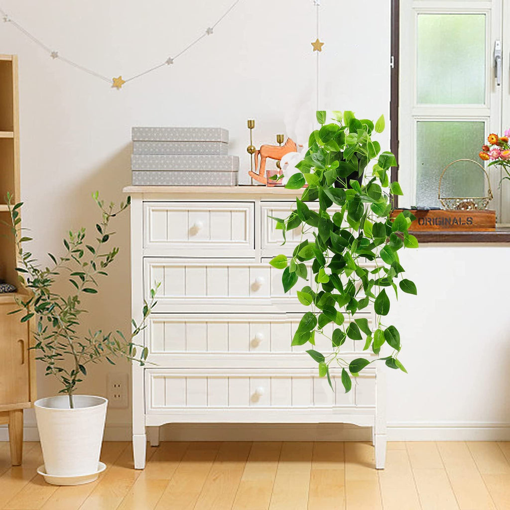 Artificial hanging ivy plant with green leaves in a black pot, displayed in a room with a white dresser and window.