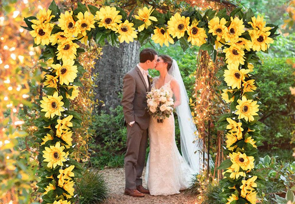 Wedding couple kissing under an archway decorated with artificial sunflower garlands and string lights.