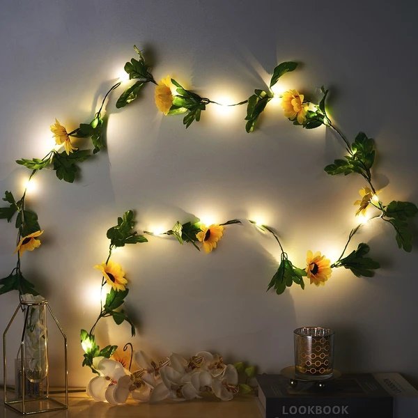 Artificial sunflower garland with LED string lights draped on a wall, with decorative items on a shelf below.