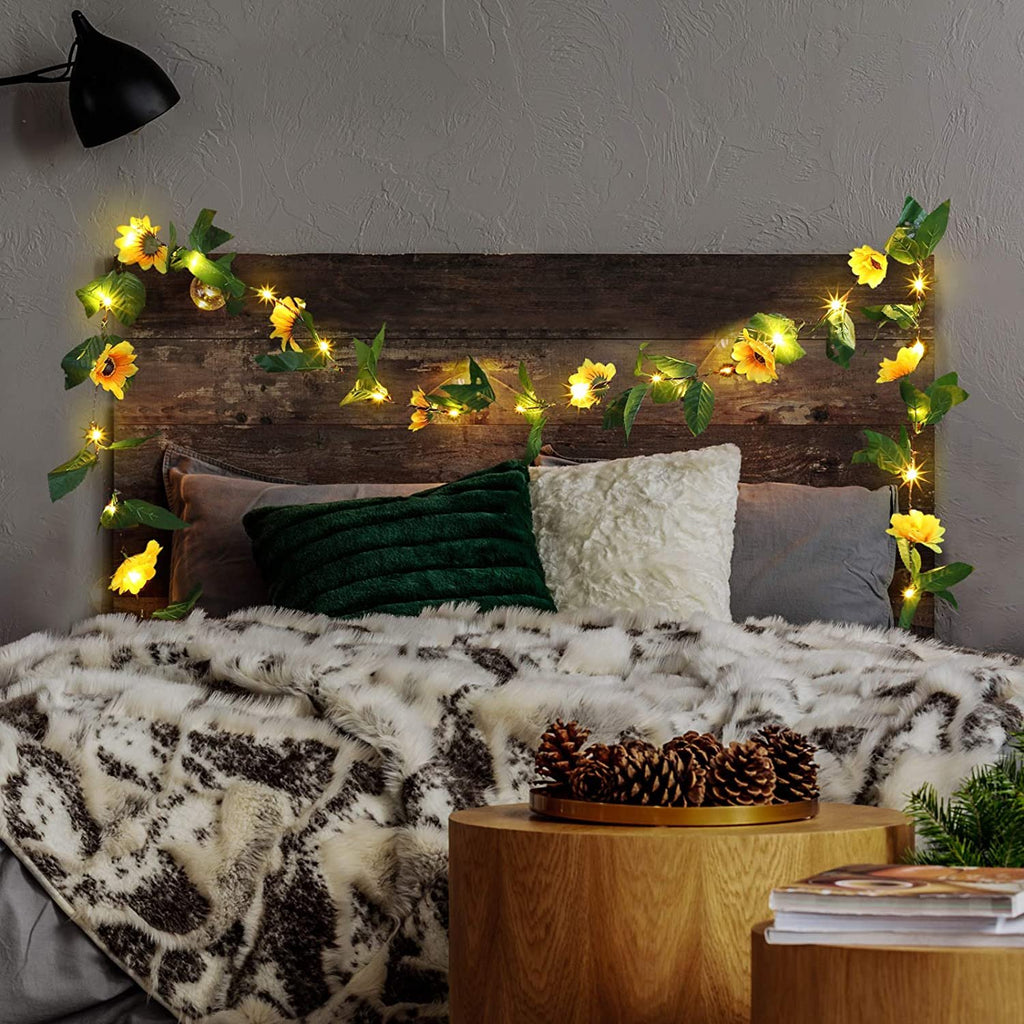 Artificial sunflower garland with LED string lights draped over a wooden headboard above a bed with pillows and a faux fur blanket.