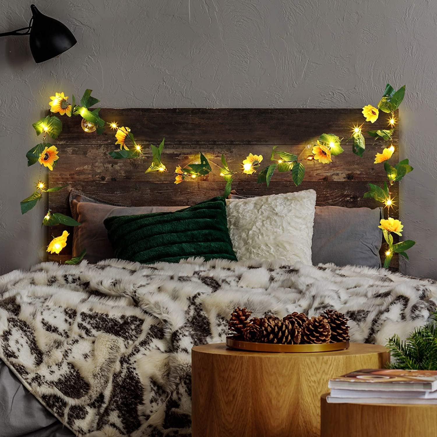 Artificial sunflower garland with LED string lights draped over a wooden headboard above a bed with pillows and a faux fur blanket.