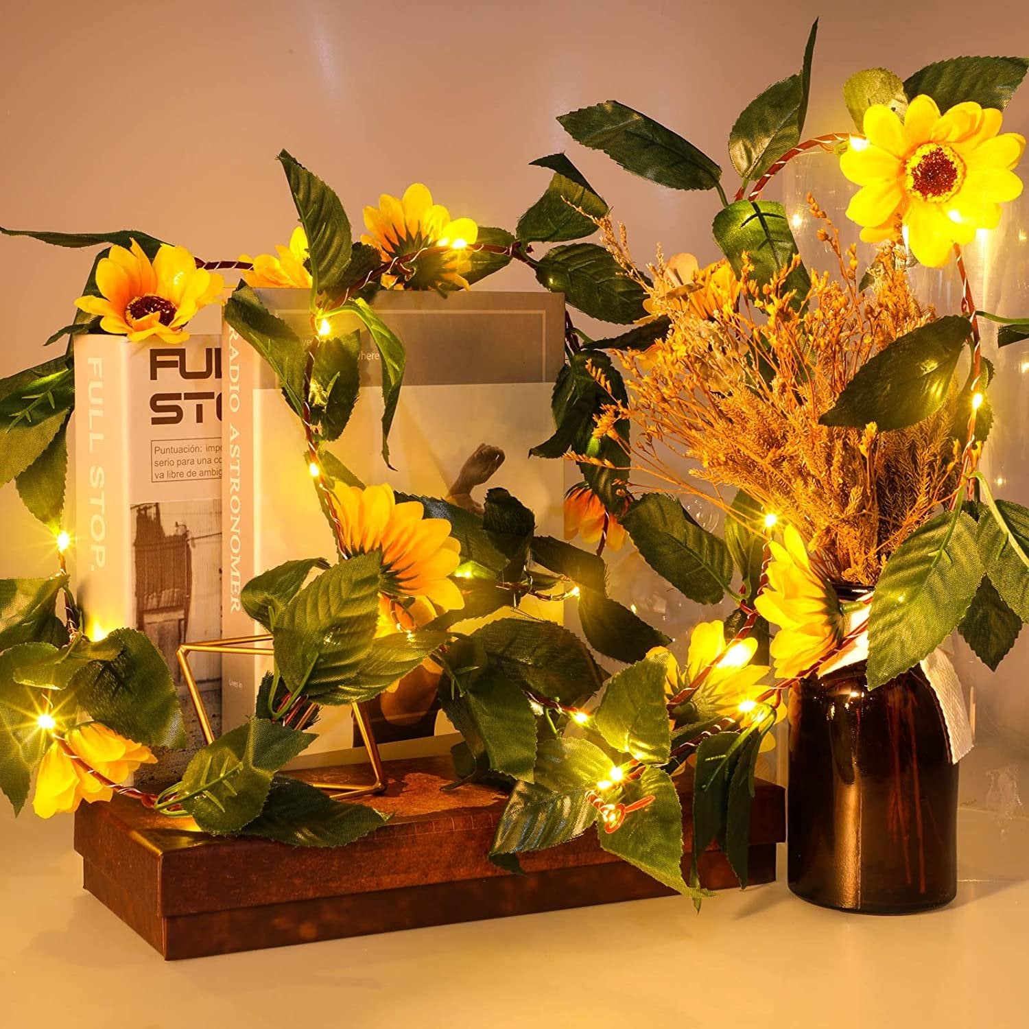 Artificial sunflower garland with LED string lights draped around books and a vase of dried flowers.