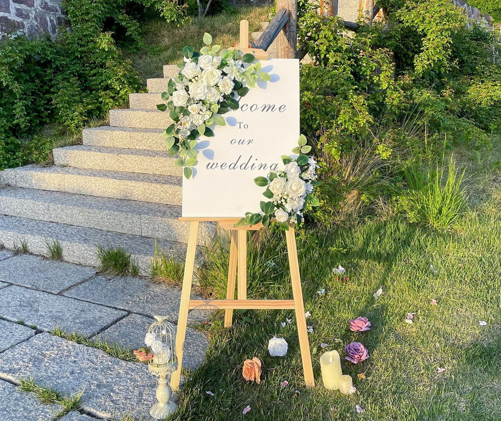 White welcome sign with floral accents and text 'Come To Our Wedding' displayed on an easel outdoors.