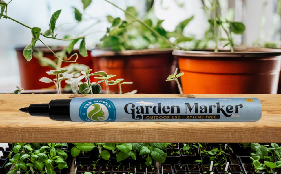 Black garden marker pen with 'Garden Marker' text and '' logo, resting on a wooden shelf with plants in pots and seed trays.