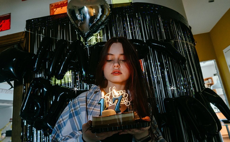 Young woman holding a birthday cake with 'Happy 17th Birthday' candles, with black tinsel curtains and balloons in the background.