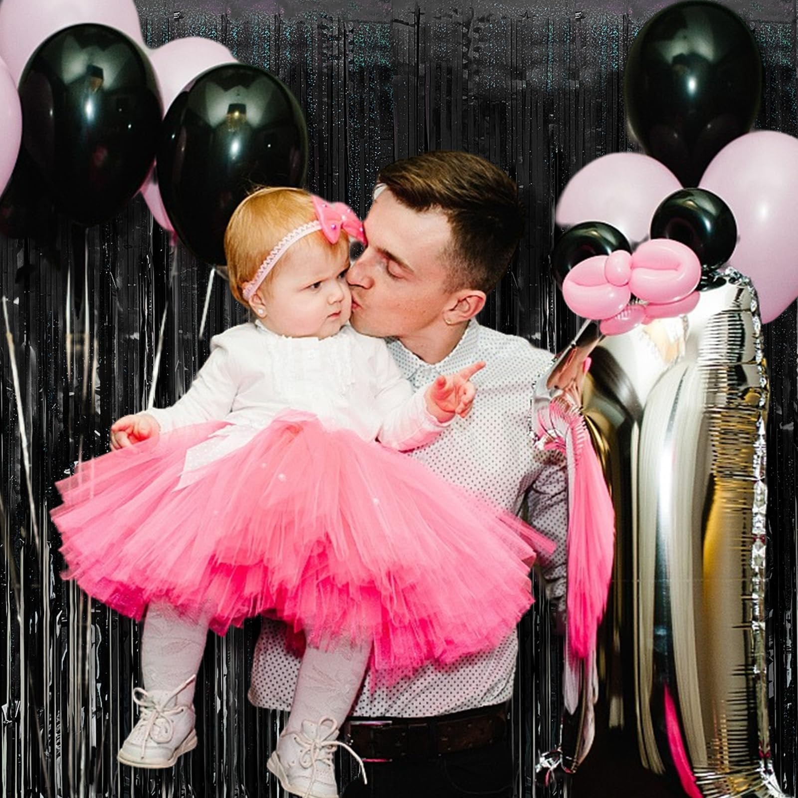 Father kissing baby daughter in pink tutu, surrounded by black and pink balloons and a silver number 1 balloon.