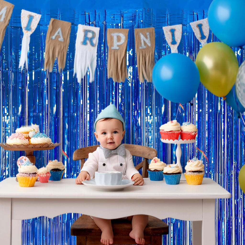 Baby sitting at a table with cupcakes and balloons, in front of a blue tinsel fringe backdrop.