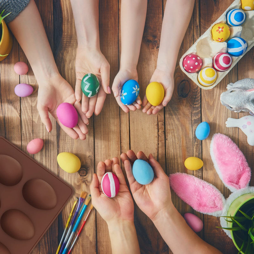 Hands holding painted Easter eggs and a brown silicone egg mold on a wooden table, with bunny ears and a plant visible.