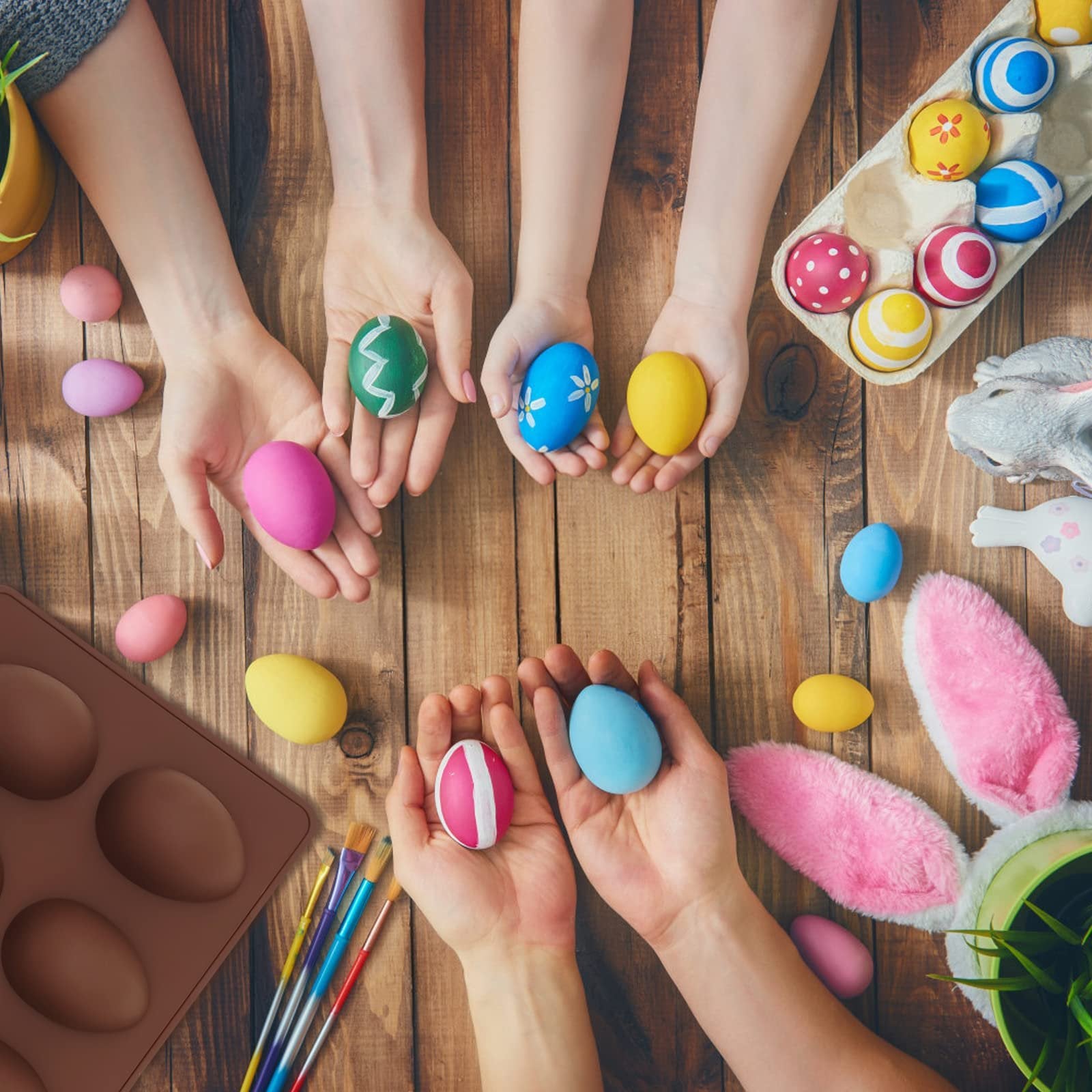 Hands holding painted Easter eggs and a brown silicone egg mold on a wooden table, with bunny ears and a plant visible.