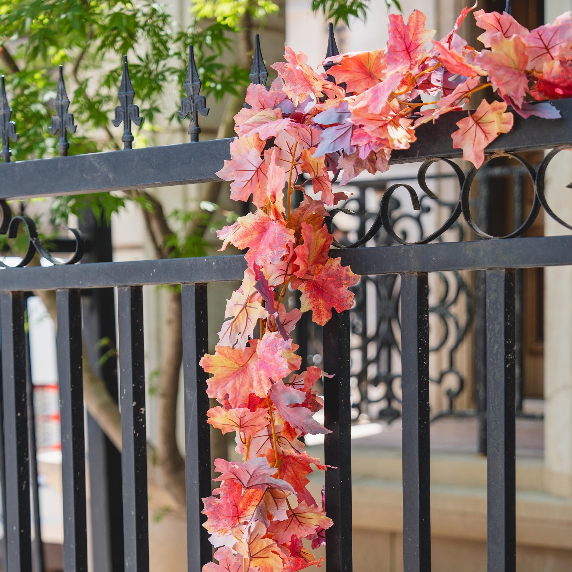 Artificial maple leaf garland in fall colors draped over a black metal fence with green foliage in the background.