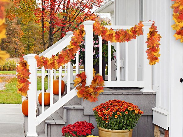 Autumn maple leaf garland draped on a white porch railing with pumpkins and mums below.