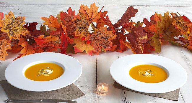 Two bowls of pumpkin soup with cream and seeds, set on a table with autumn leaf garland and a lit candle.