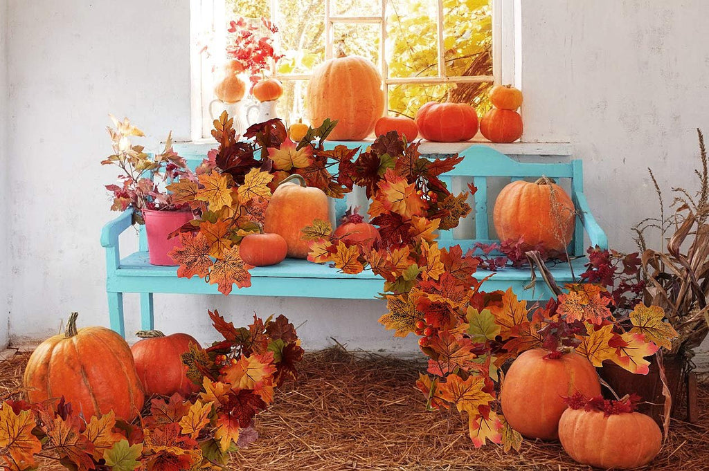 Autumnal scene with pumpkins and maple leaf garlands arranged on and around a blue bench, with a window in the background.