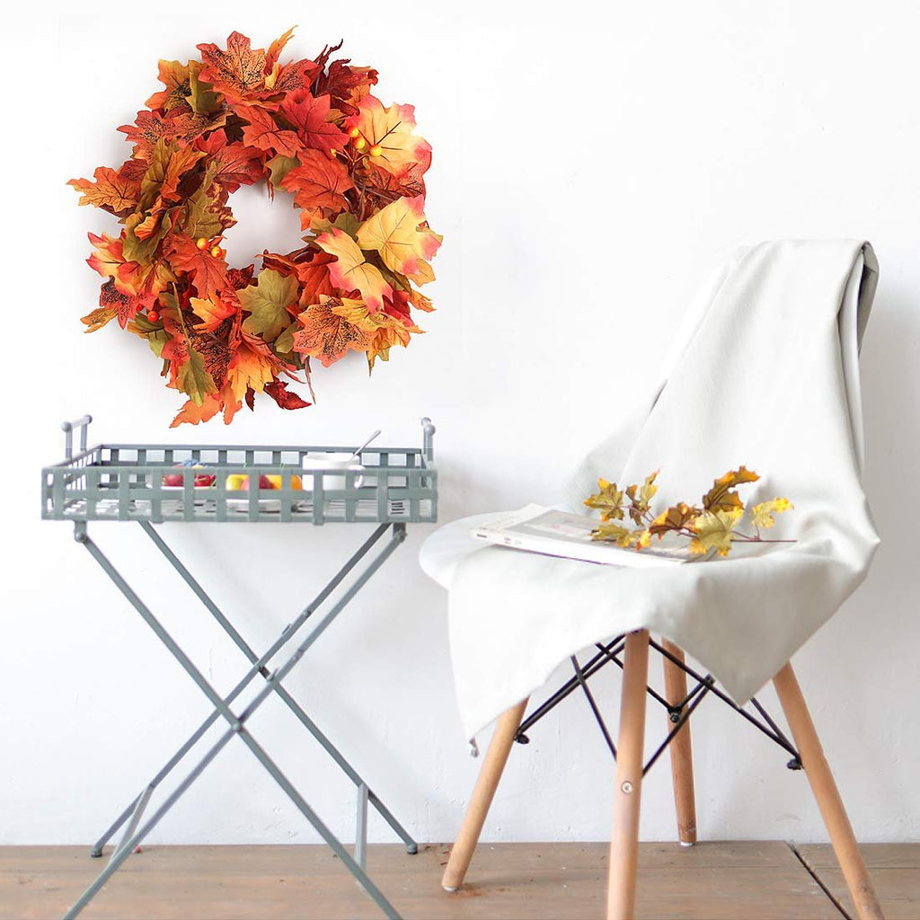 Autumn maple leaf wreath hanging on a white wall above a metal tray table and a chair draped with a white cloth.