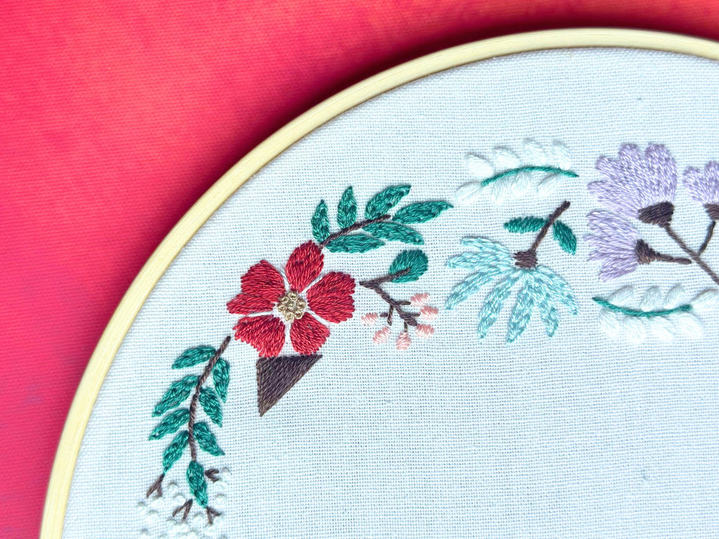 Close-up of a floral embroidery design on white fabric within a bamboo hoop, set against a pink background.