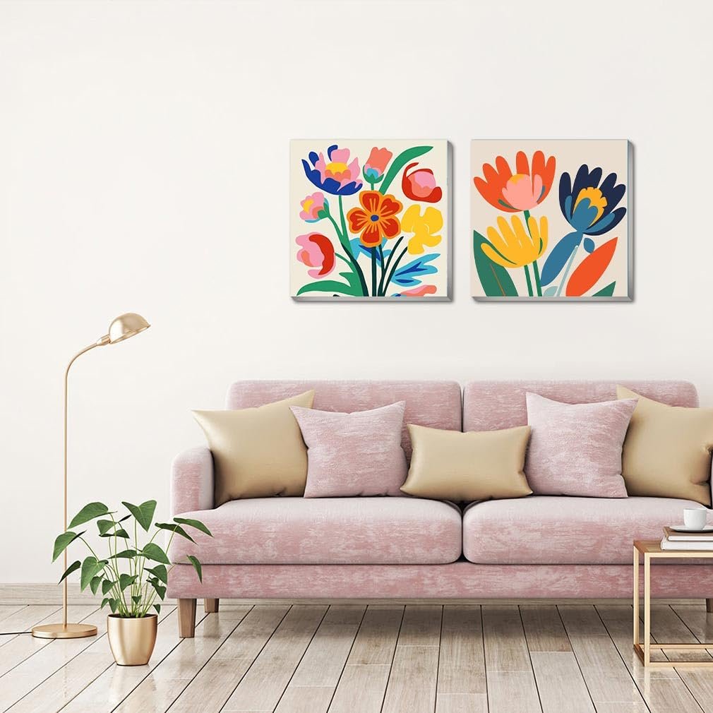 Two framed abstract floral paintings hanging above a pink sofa with gold and pink pillows, next to a floor lamp and potted plant.