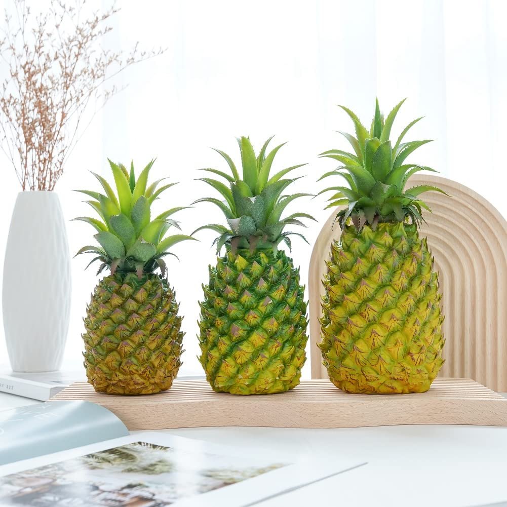 Three artificial pineapples of varying sizes displayed on a wooden stand with a vase of dried flowers in the background.