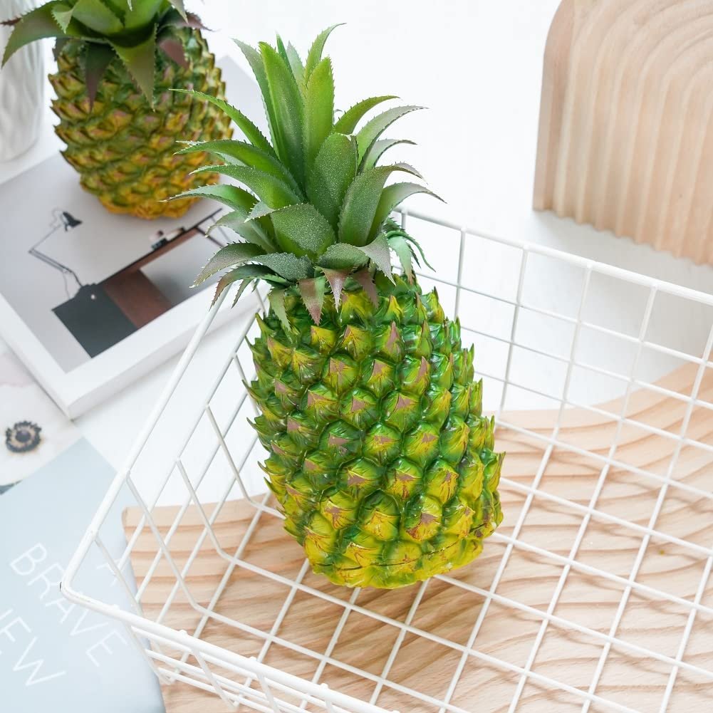 Two artificial green pineapples in a white wire basket on a wooden tray, with a book and wooden decor in the background.