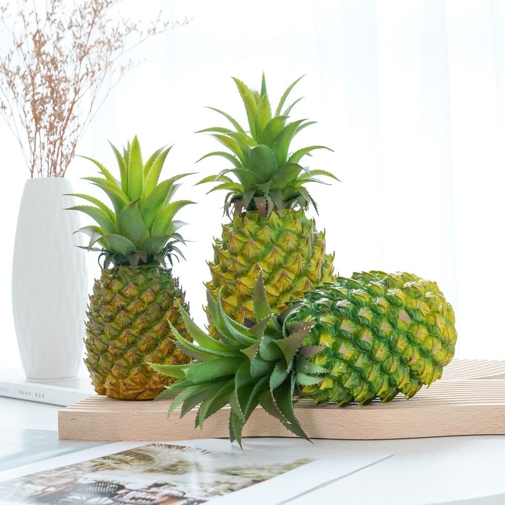 Three artificial pineapples, two upright and one lying down, on a wooden board with a vase of dried flowers in the background.