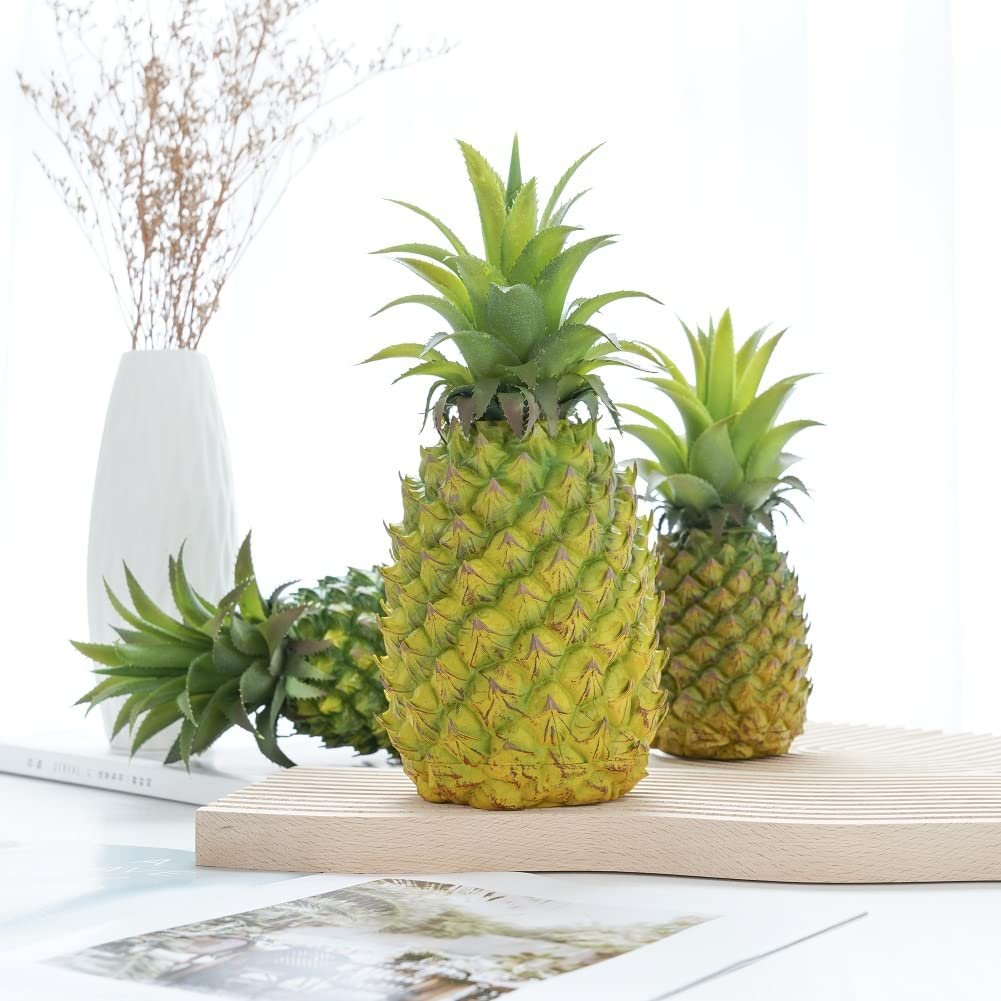 Two artificial green pineapples on a wooden stand with a vase of dried flowers in the background.