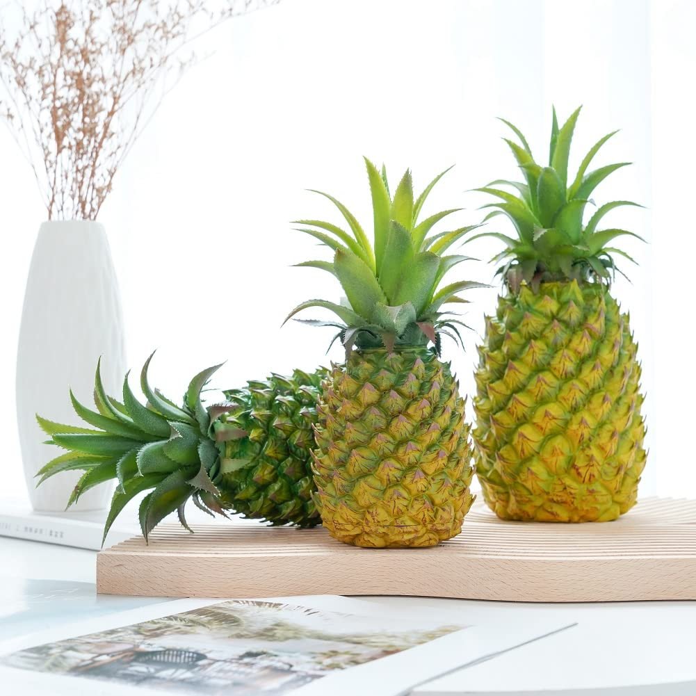 Two green artificial pineapples on a wooden board with a white vase and dried flowers in the background.