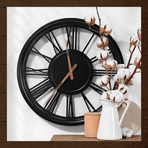Black wall clock with Roman numerals and wood hands, next to a white pitcher with cotton branches.