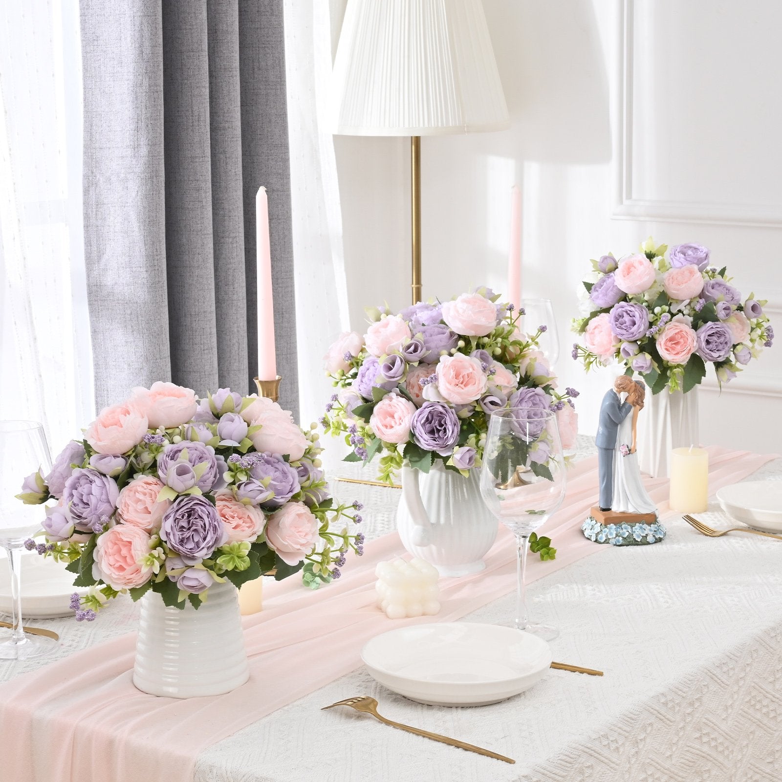 Three bouquets of pink and purple artificial peonies arranged on a table with candles and place settings.