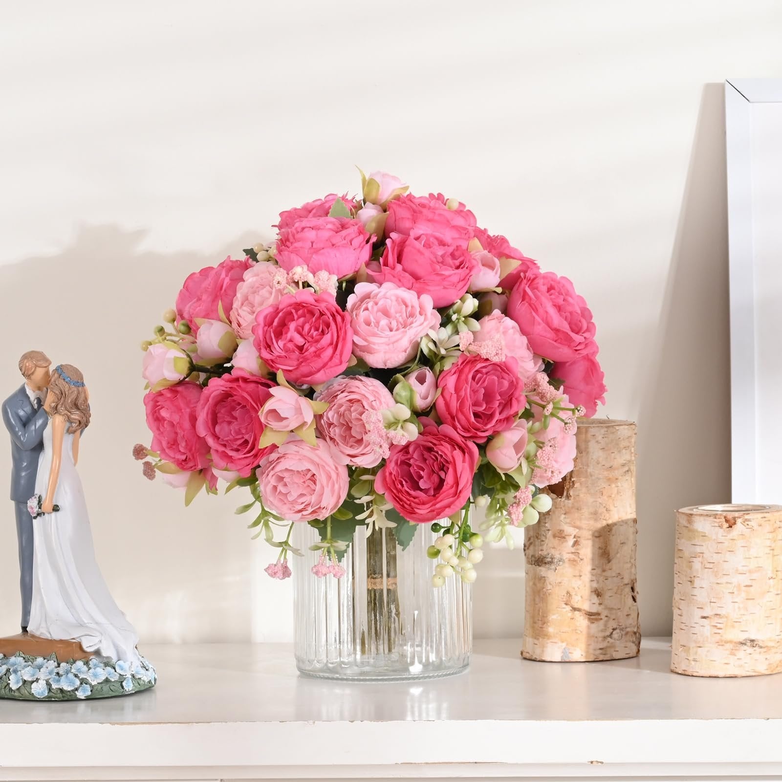 Two bouquets of pink silk peonies in a clear glass vase, with a wedding cake topper and birch log candle holders on a shelf.