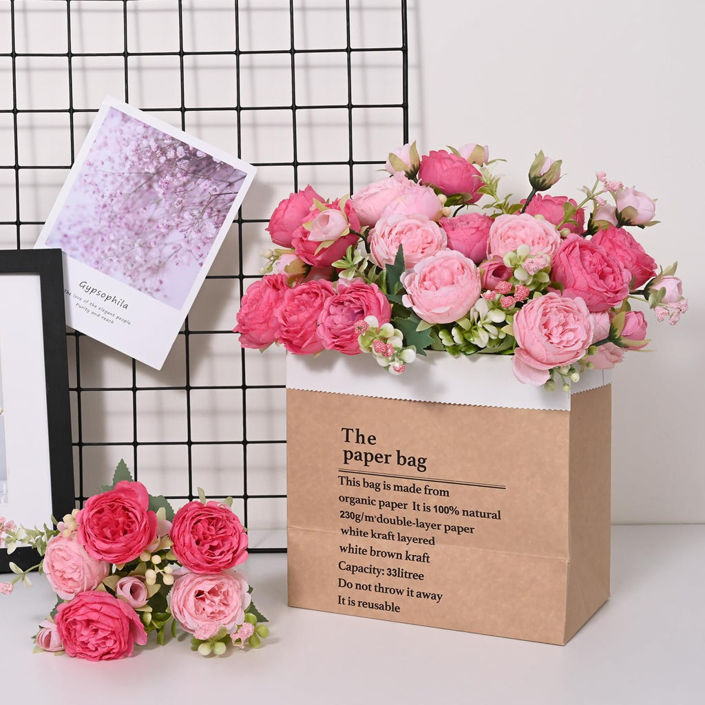 Two bouquets of pink artificial peonies arranged in a brown paper bag and a small bunch on a white surface, with a framed picture and a grid background.