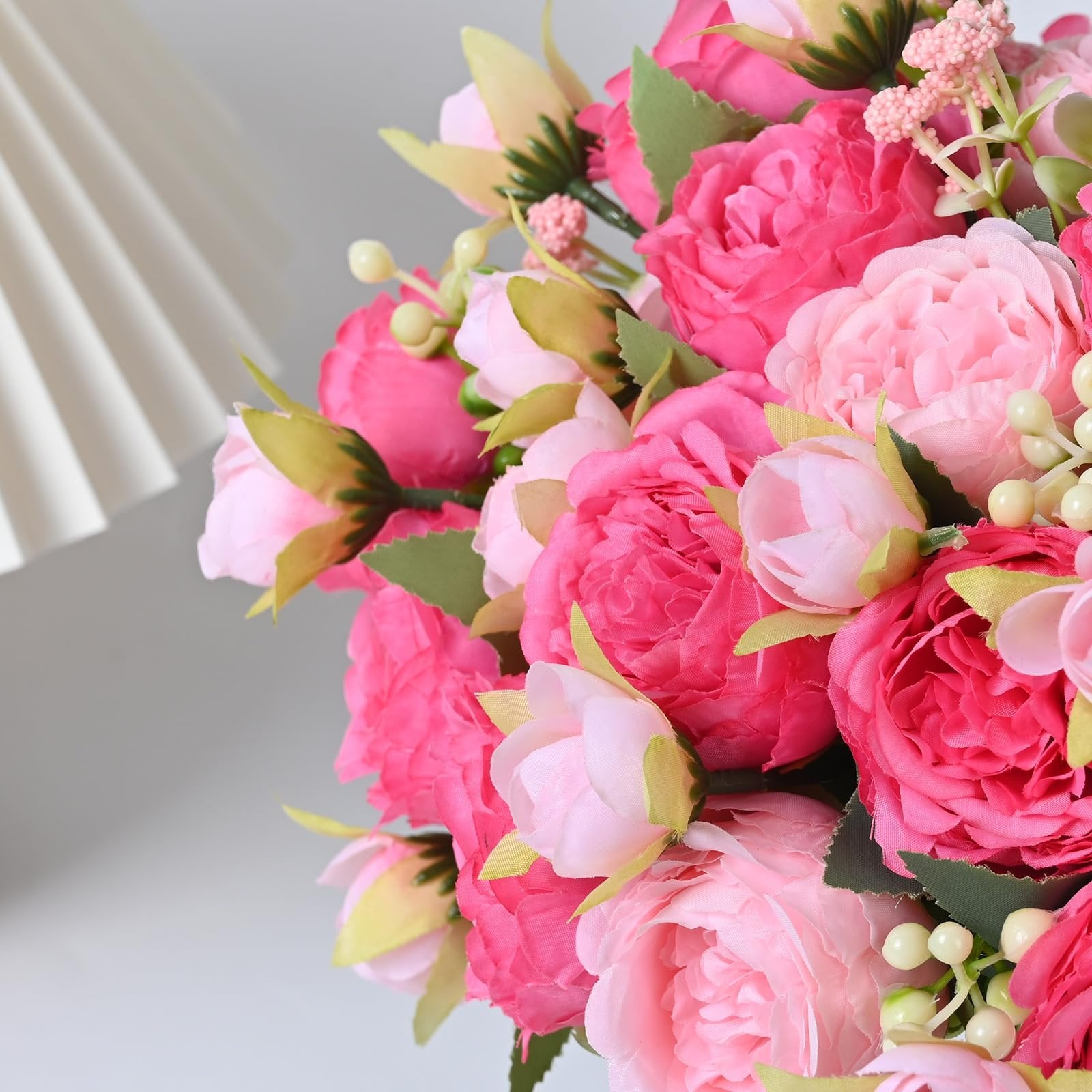 Close-up of a bouquet of mixed pink silk peonies with buds and green leaves, set against a white background with a pleated lamp shade.