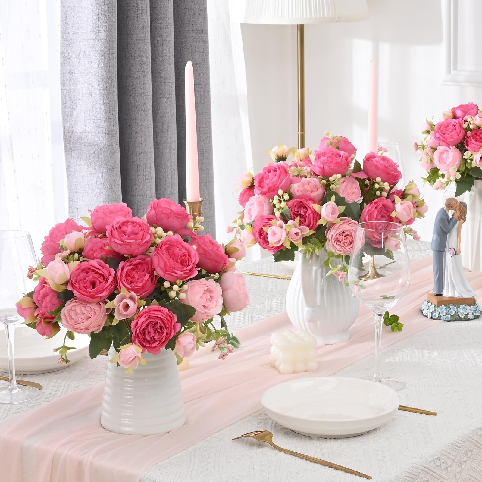 Two bouquets of pink silk peonies in white vases on a table setting with candles and a wedding cake topper.