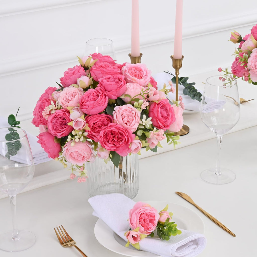 Table setting with a bouquet of pink peonies in a vase, wine glasses, and napkins with floral accents.