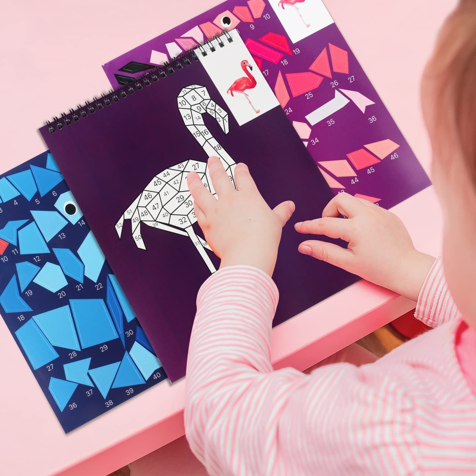 Child's hands working on a paint by number sticker book featuring a flamingo design with numbered pieces.