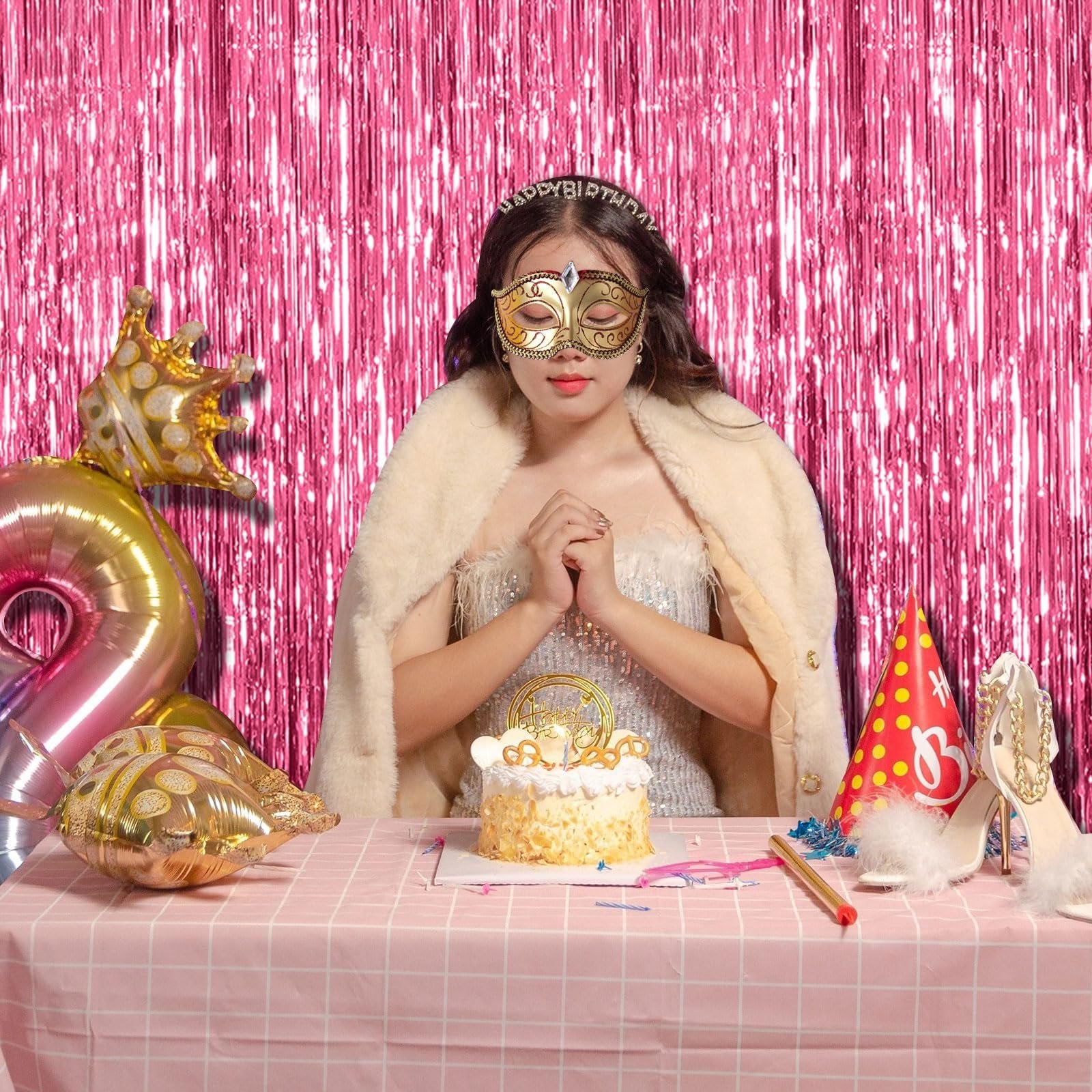 Woman wearing a mask and crown sits at a table with a cake, balloons, and party hat, in front of a pink tinsel backdrop.