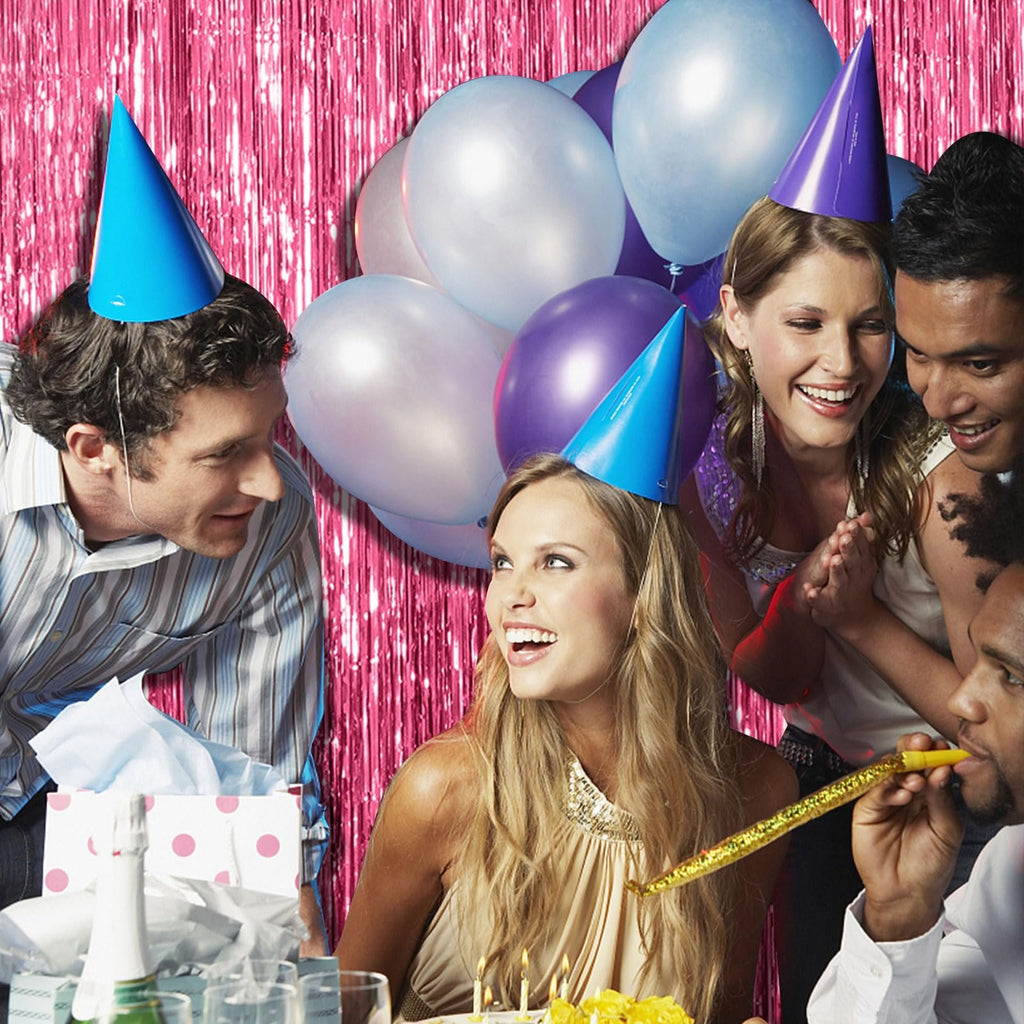 Group of people celebrating with party hats and balloons, with a pink tinsel backdrop.