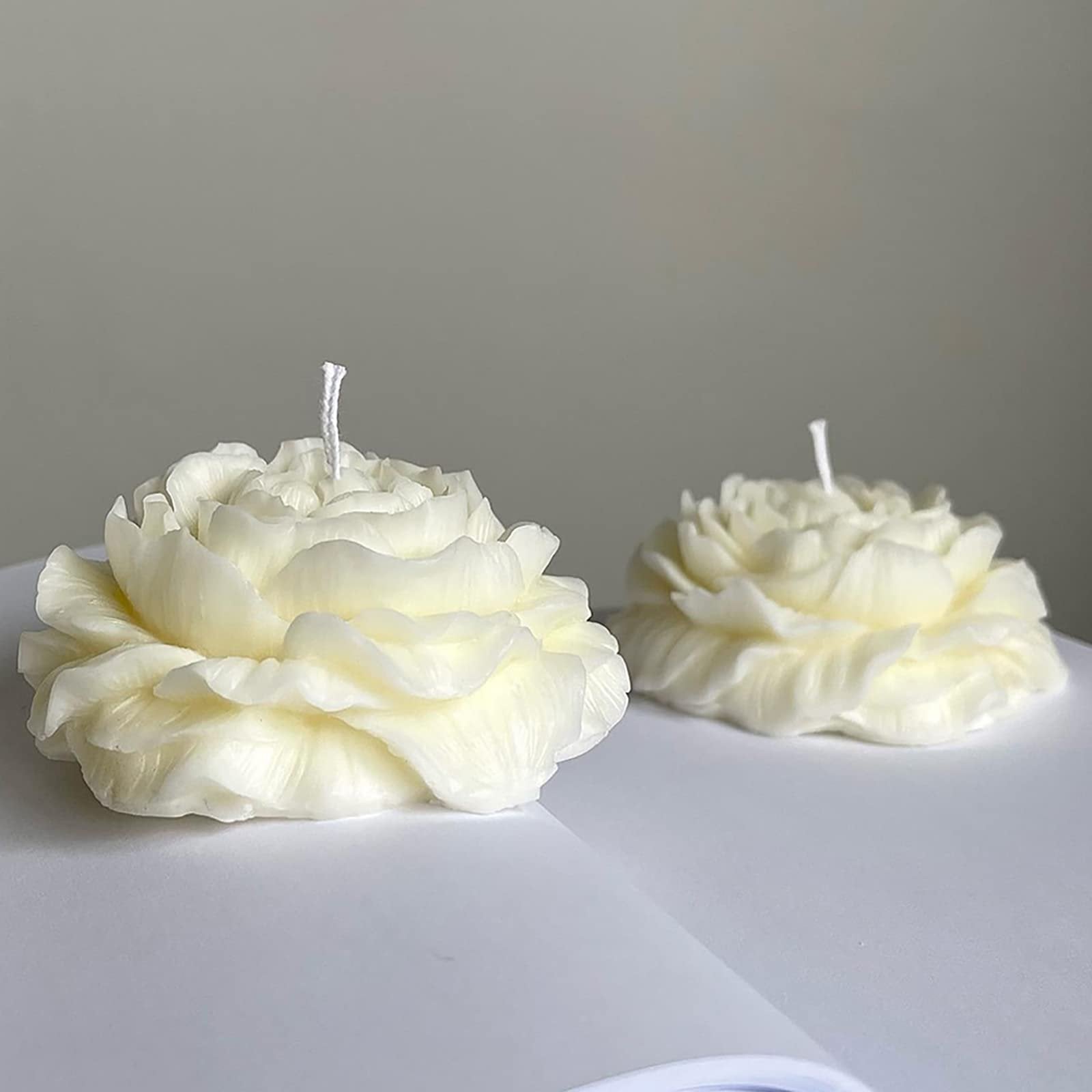 Two white peony-shaped candles with wicks on a white surface against a grey background.