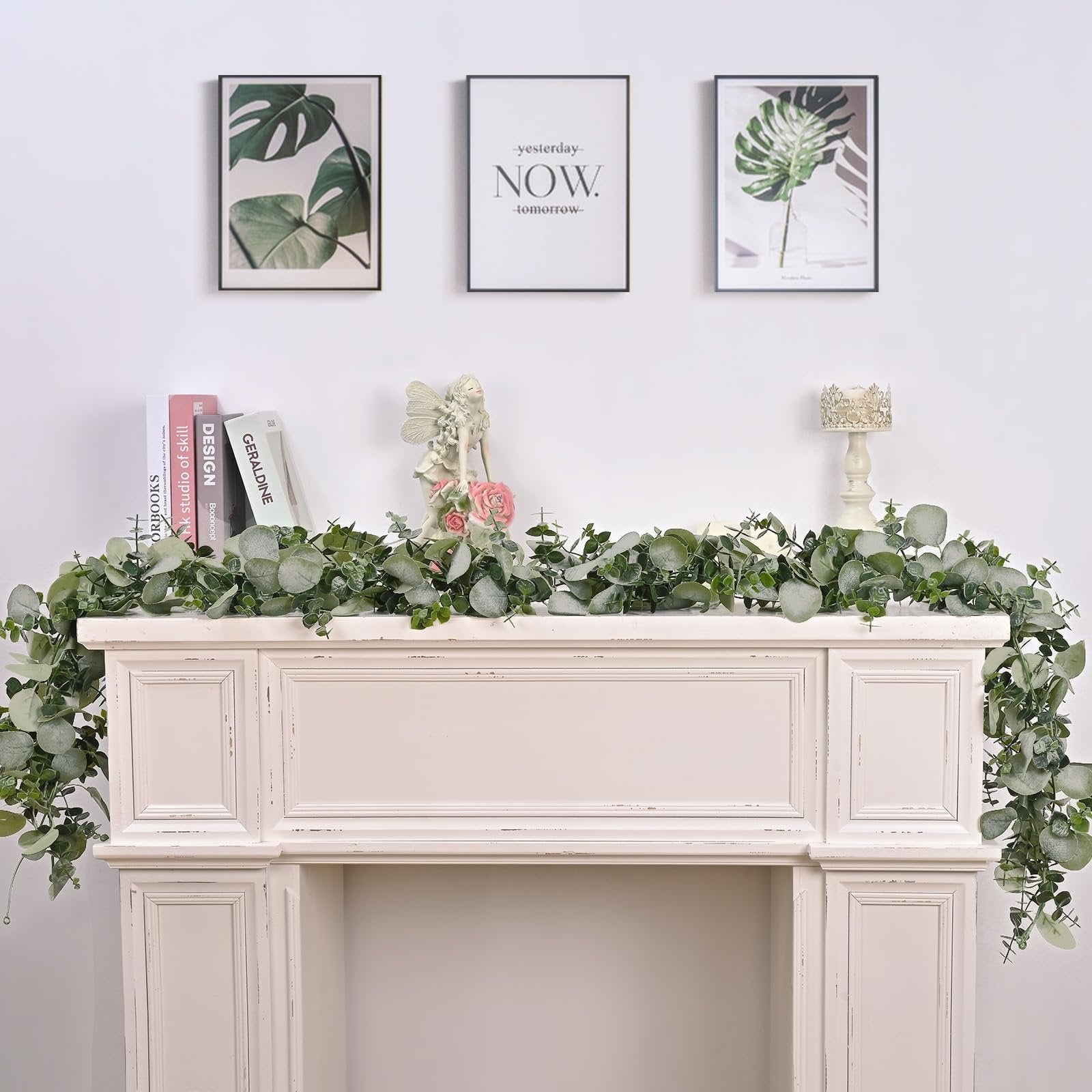 Artificial eucalyptus garland draped over a white mantelpiece with framed art and decorative items above.