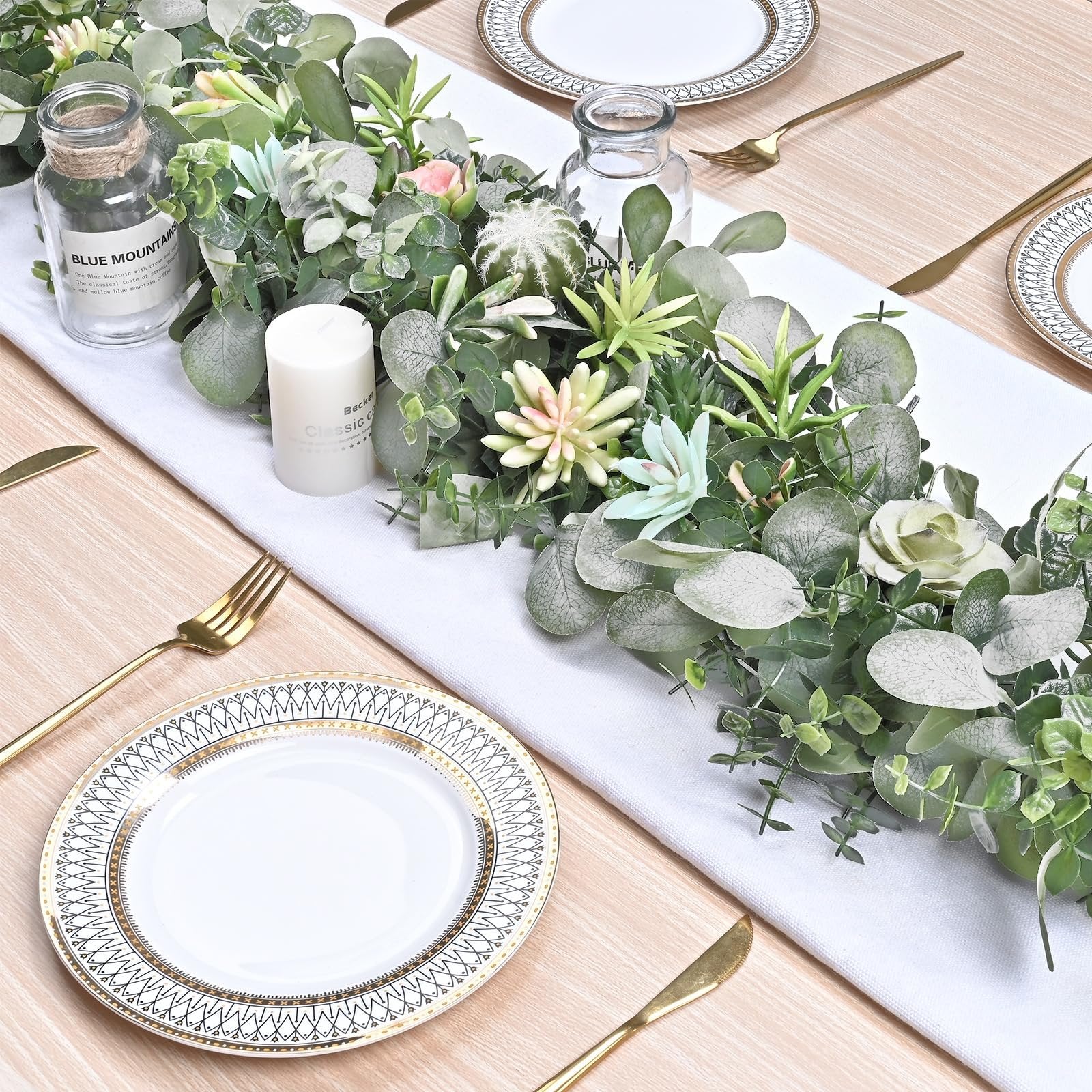 Artificial eucalyptus garland with succulents and a candle on a white table runner, set with plates and gold cutlery.