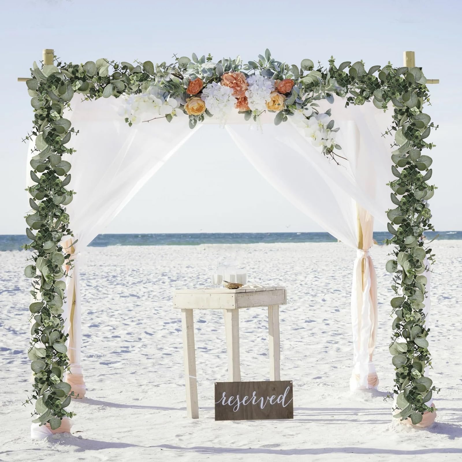 Beach wedding arch decorated with artificial eucalyptus garland and flowers, with a small table and 'reserved' sign on the sand.