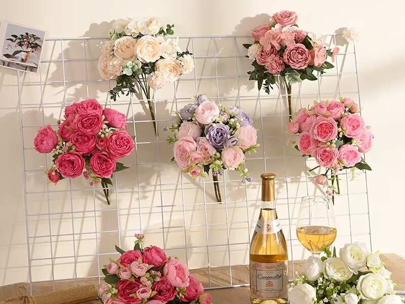 Assortment of artificial peony flower bouquets in various colors displayed on a white grid wall, with a bottle of wine and a glass nearby.