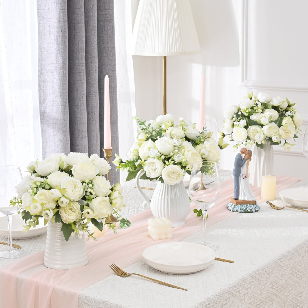 Table setting with white artificial peony flower bouquets in vases, candles, and a wedding couple figurine.