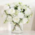 Two bouquets of white artificial peonies and buds in a clear ribbed glass vase on a white surface.
