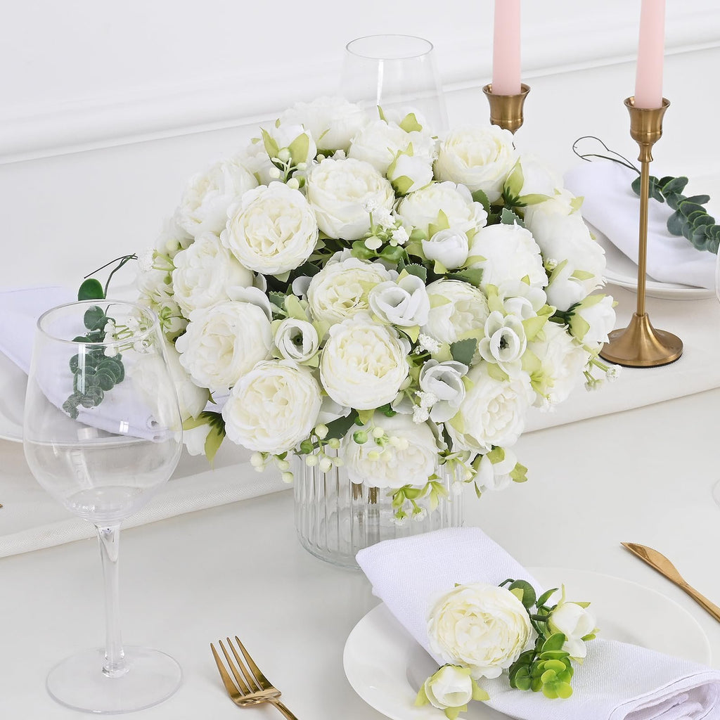 White artificial peony flower bouquet centerpiece on a table setting with gold cutlery and pink candles.