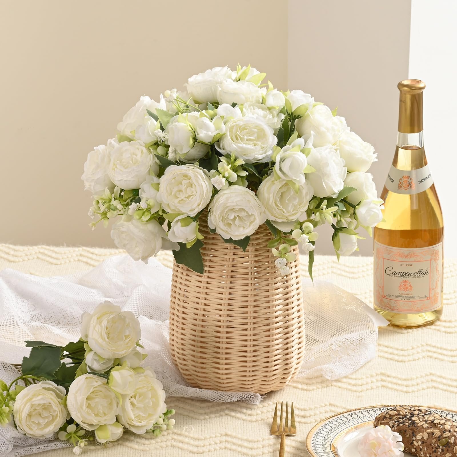 White silk peony flower bouquet in a wicker vase with a bottle of ice wine and bread on a table