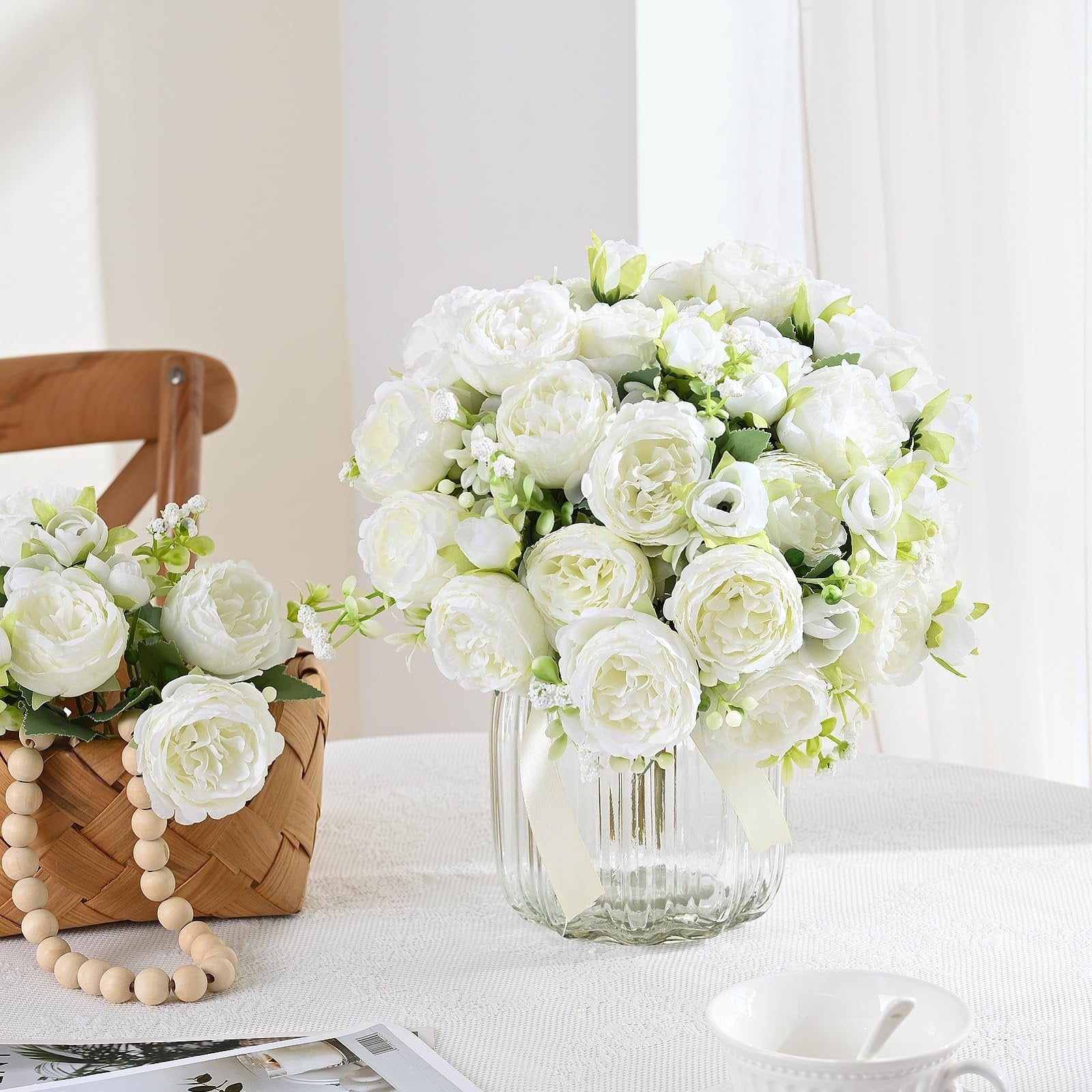 Two bouquets of white artificial peonies, one in a glass vase and one in a basket, on a white tablecloth.