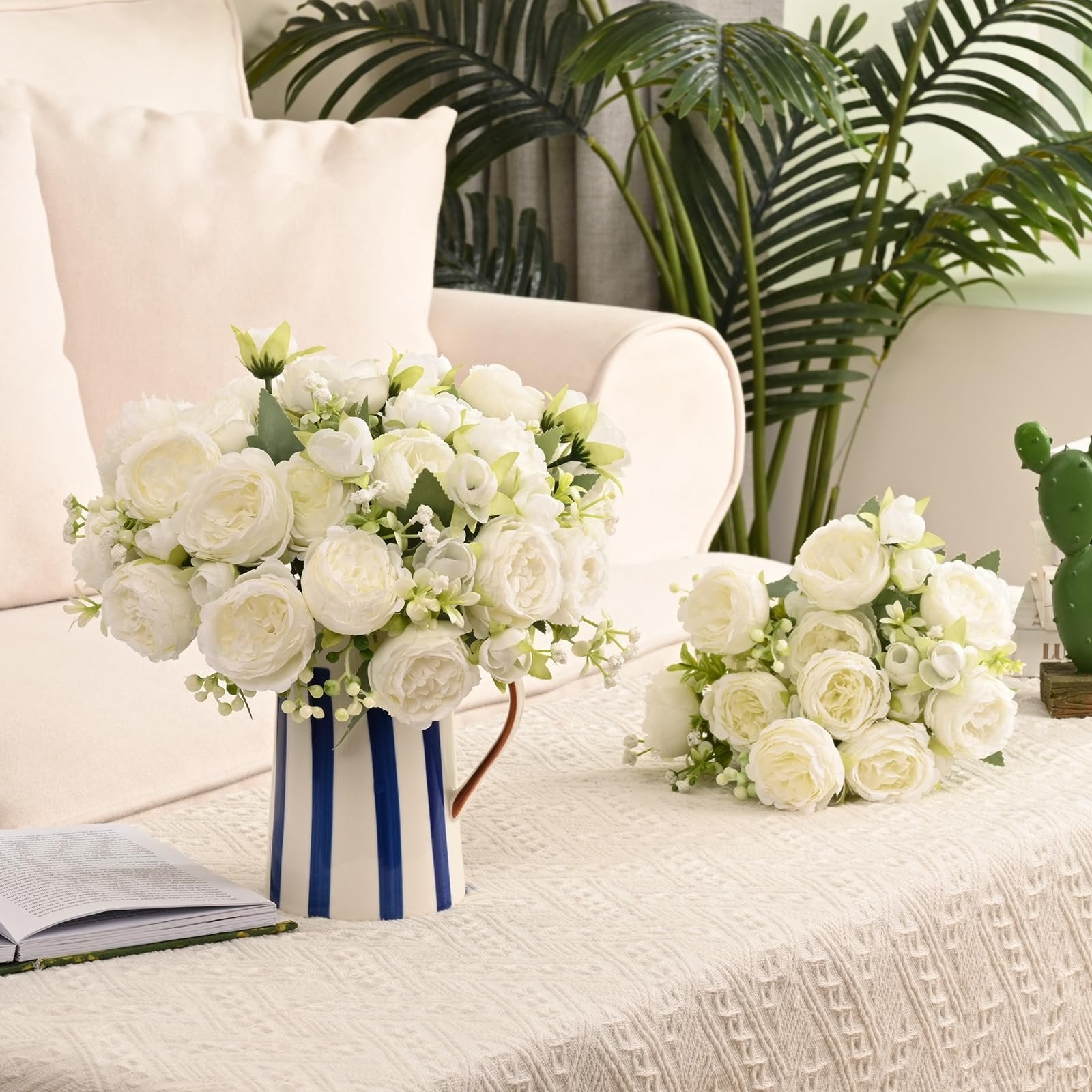 Two bouquets of white artificial peonies with buds and greenery, one in a blue and white striped vase, on a textured tablecloth.