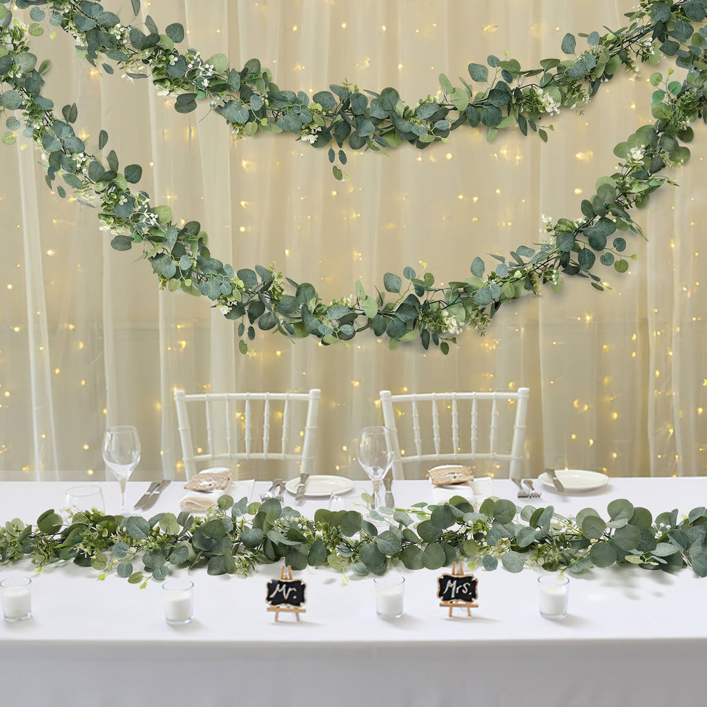 Artificial eucalyptus garland with white flowers draped on a table and wall with fairy lights.