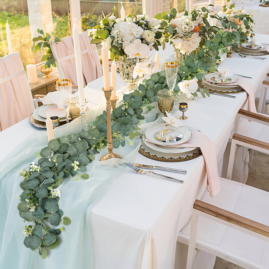Artificial eucalyptus garland with white flowers draped across a formal dining table setting with place settings and candles.